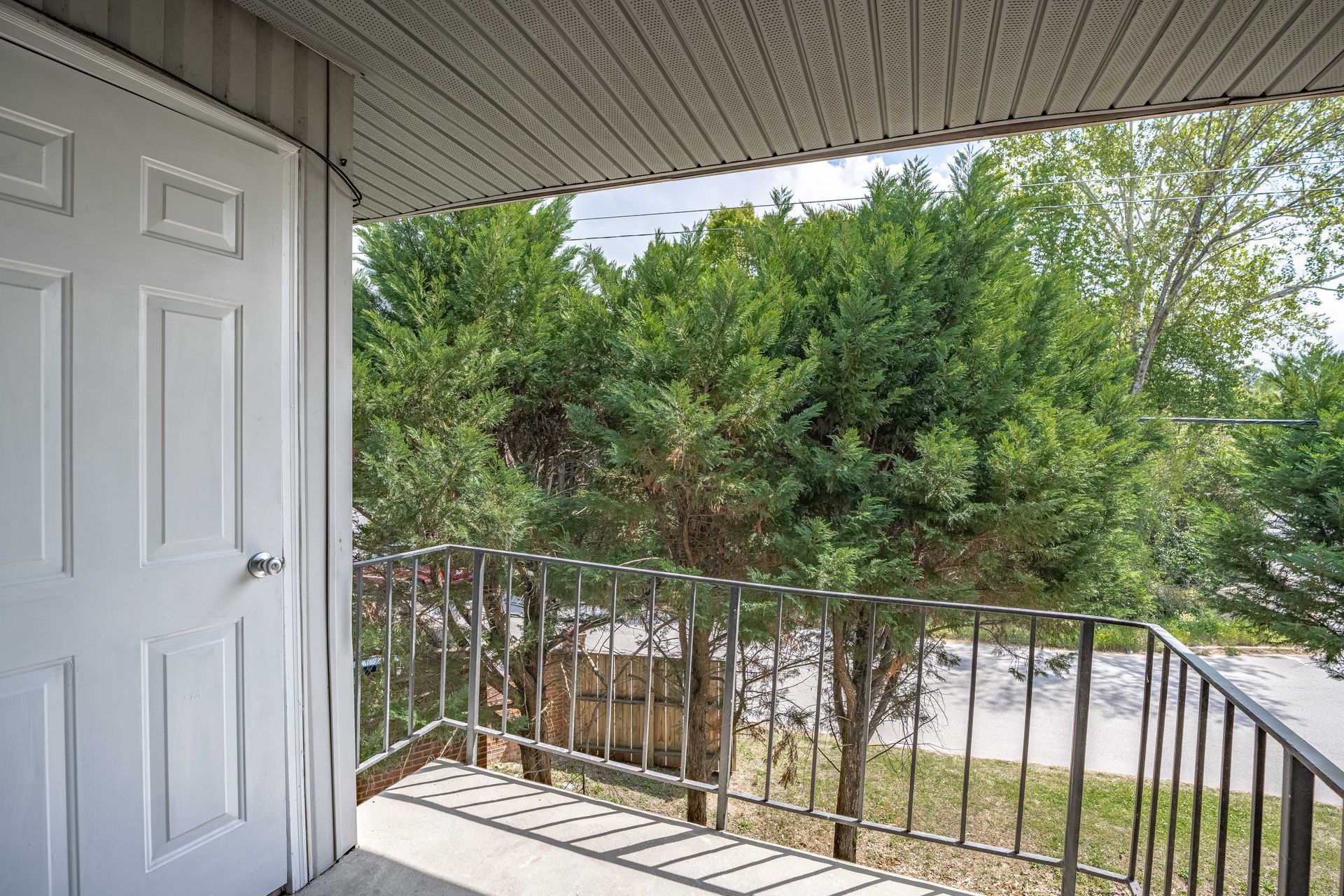 A white door leads to an outdoor balcony with a metal railing, overlooking green trees and a road.