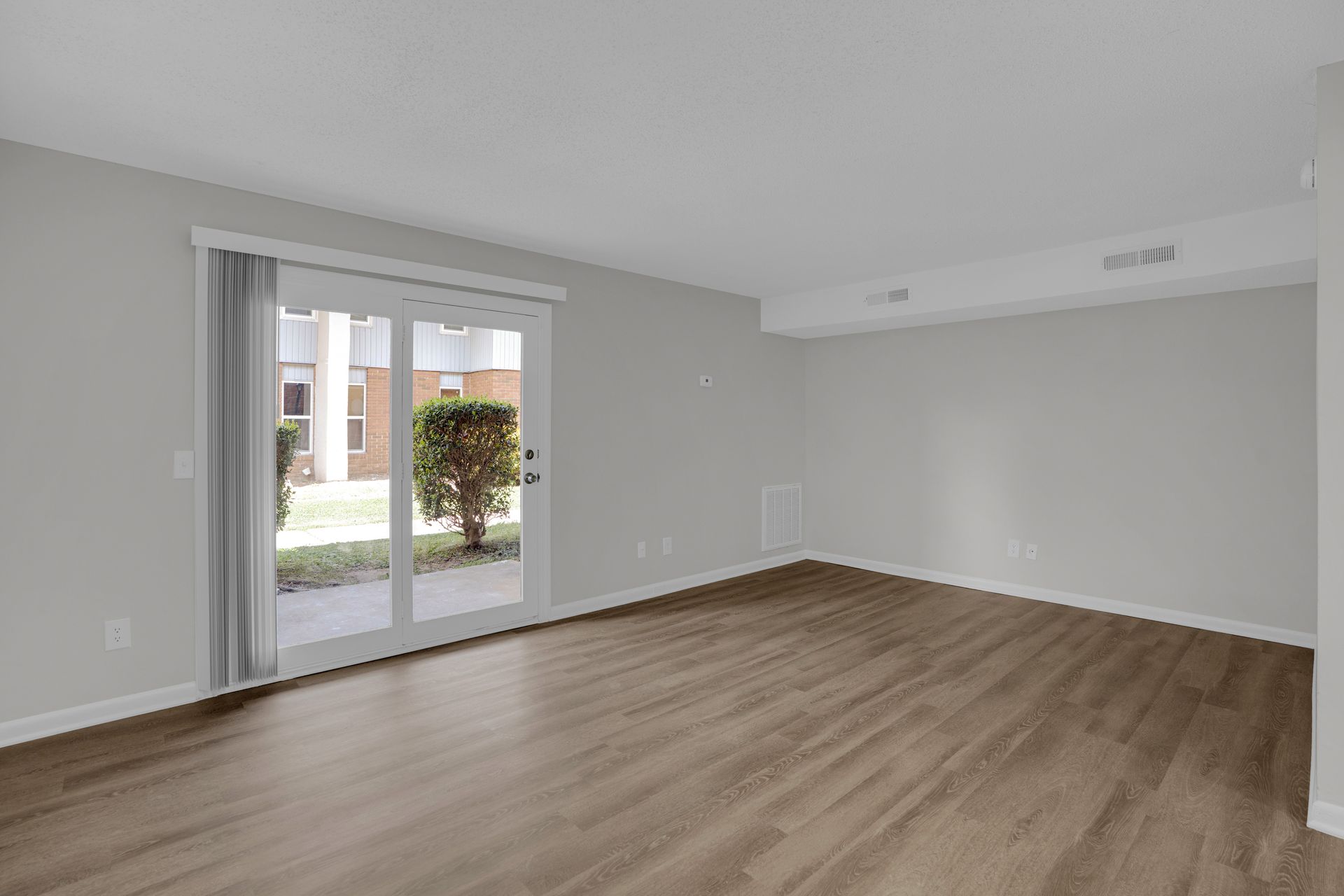 An empty, unfurnished living area with light gray walls, wood-look flooring, and a glass sliding door leading outdoors.