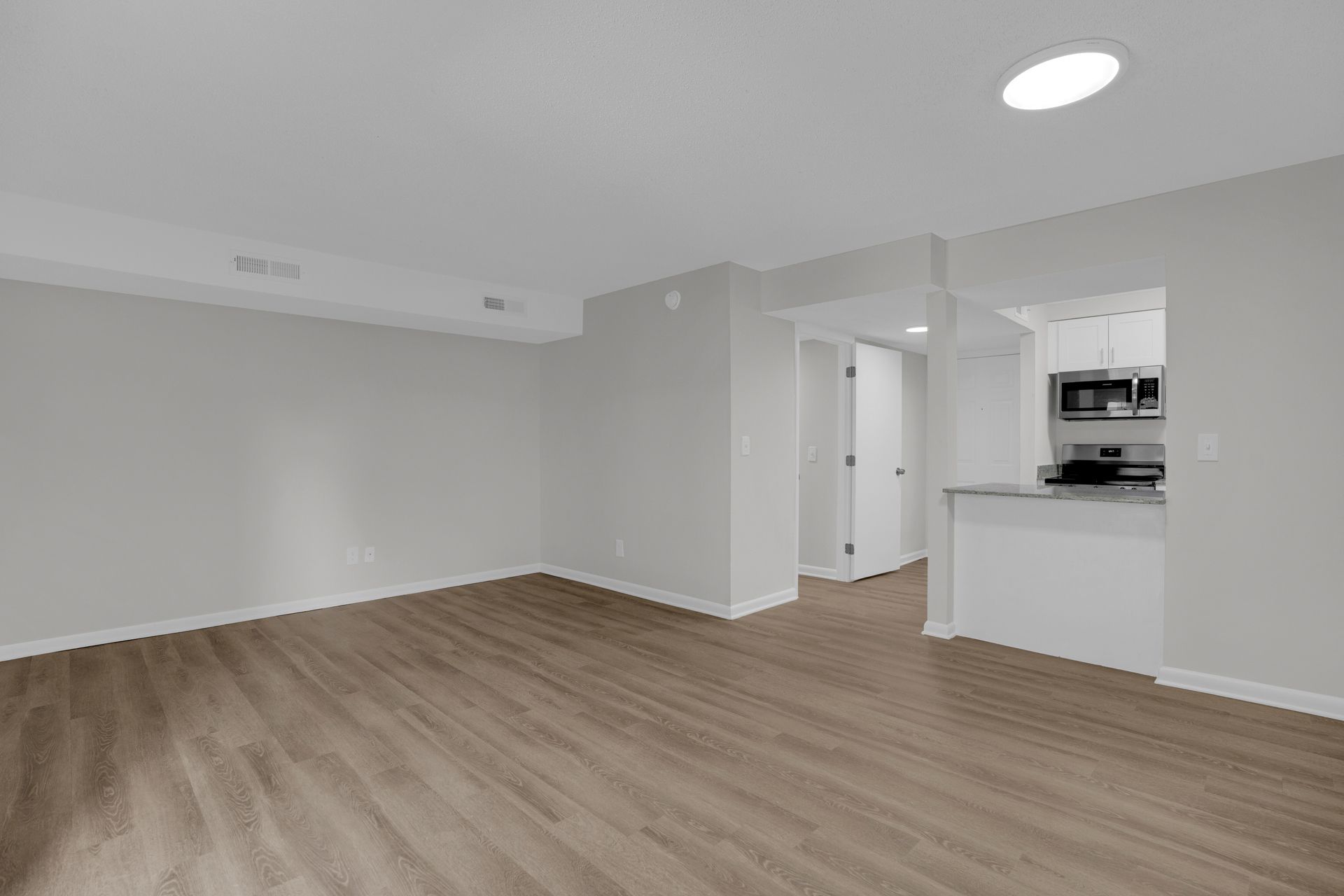 A bright, empty living room with light wood-style flooring, gray walls, and an open doorway leading to a kitchen.