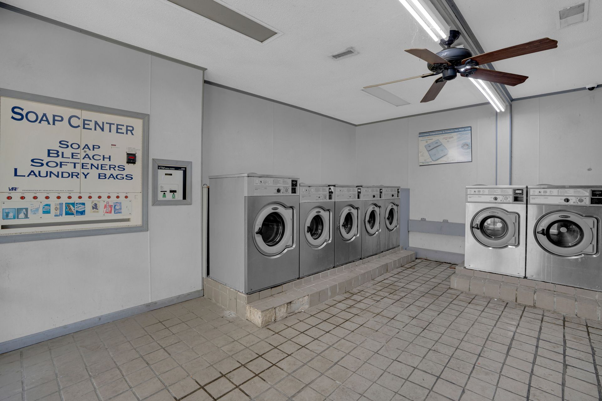 A laundry room with a row of five stainless steel washing machines on a raised platform, tile floor, and a soap dispenser.