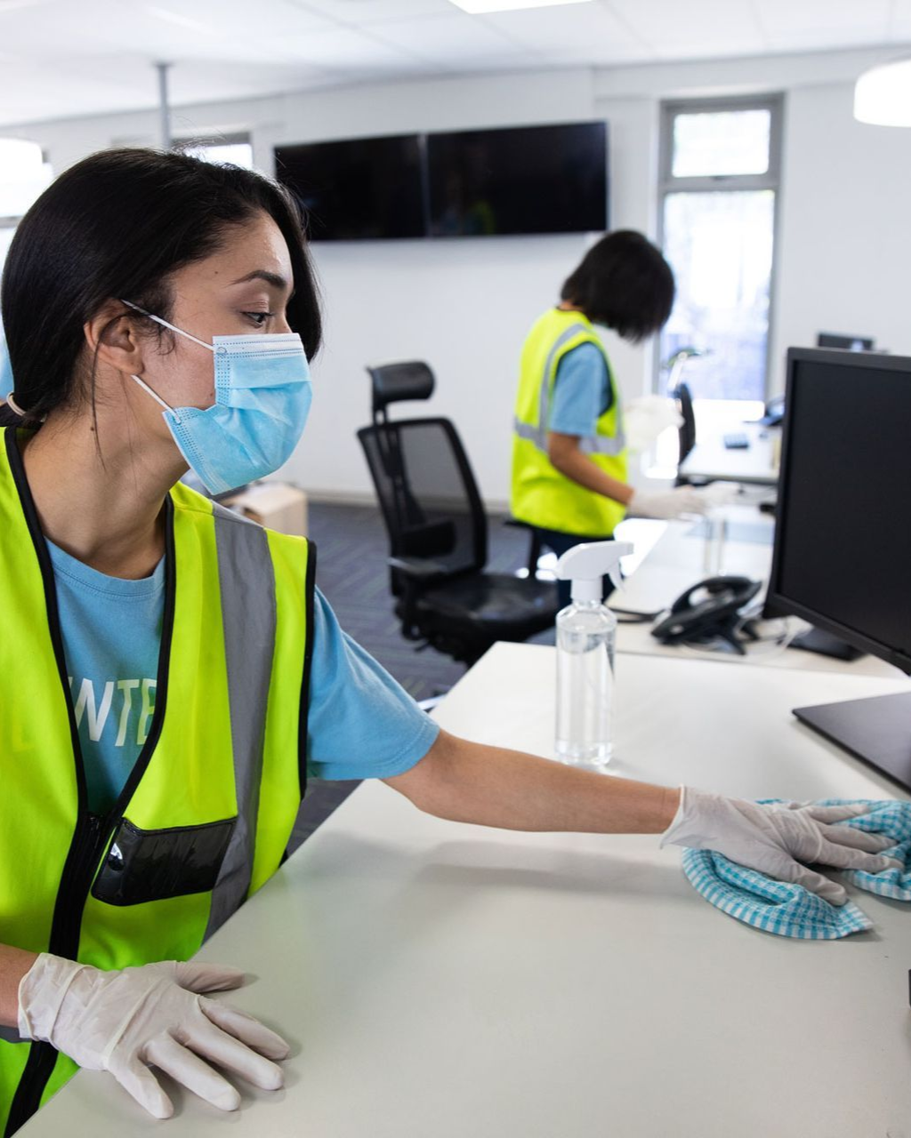 Person in mask and gloves wiping a desk in an office setting; another person cleans in the background.