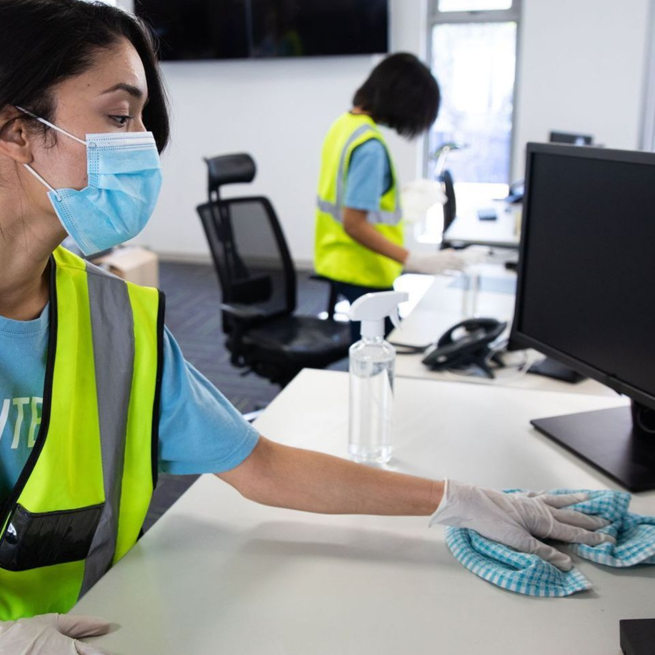 Two cleaning staff in yellow high-visibility vests, masks, and gloves wipe down office desks and computer monitors.