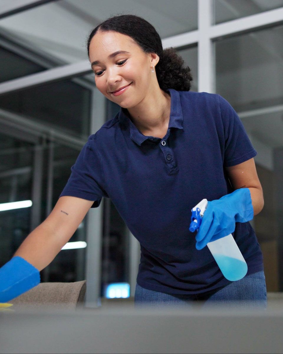 Person wearing blue gloves, cleaning a surface with a spray bottle in an office setting.