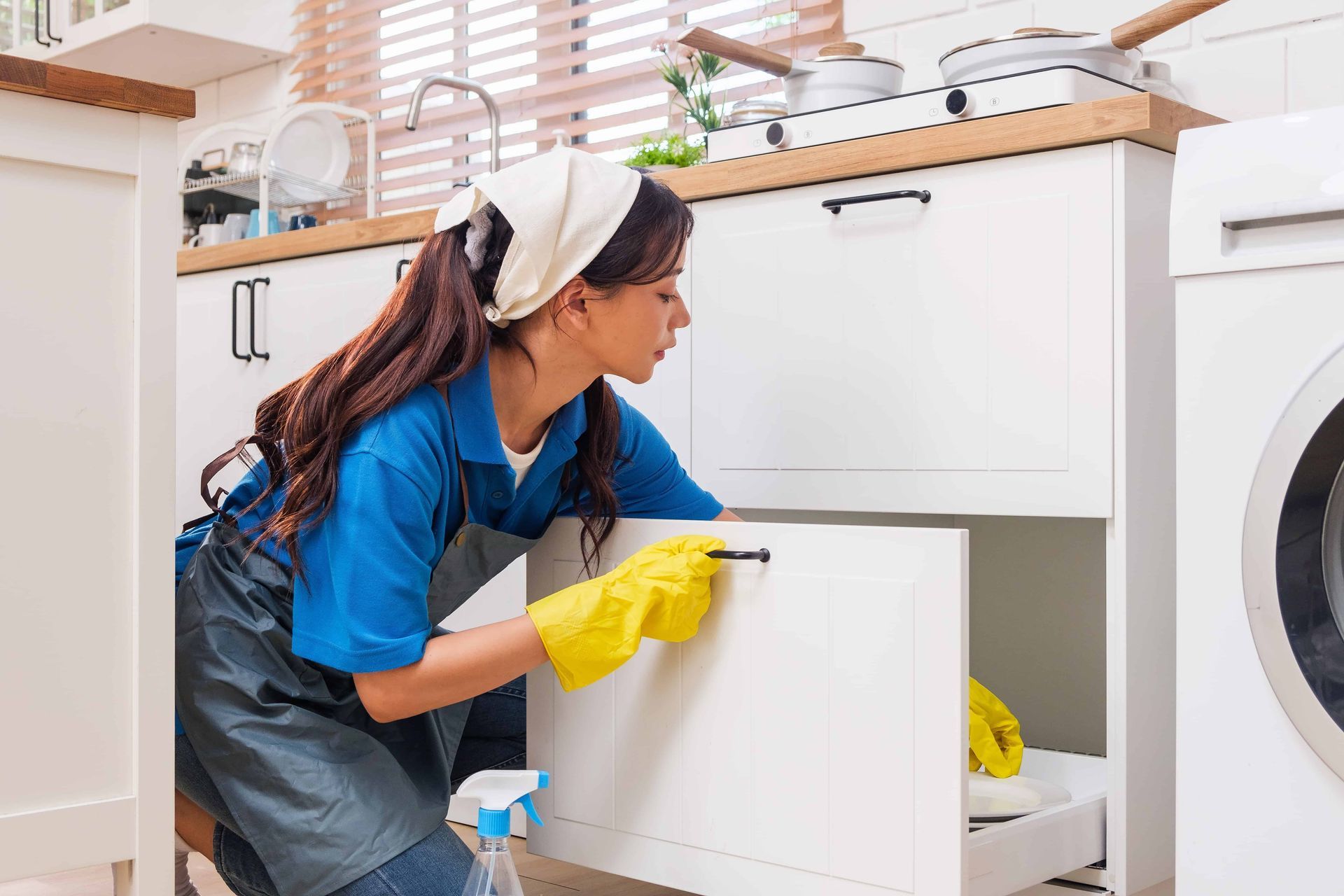 Cleaner in blue shirt and yellow gloves wiping a kitchen cabinet open drawer.