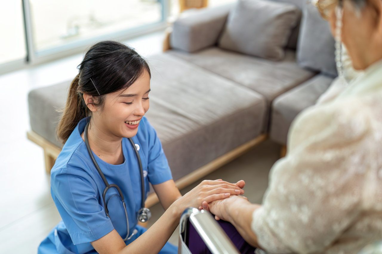 A smiling medical professional in blue scrubs kneels to hold the hands of a person sitting in a wheelchair.