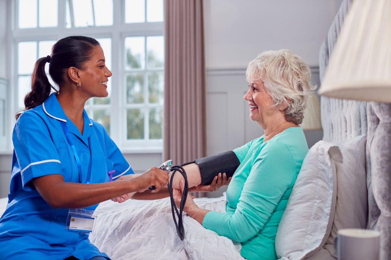 A healthcare worker in a blue uniform takes a blood pressure reading of a patient sitting on a bed in a bright room.