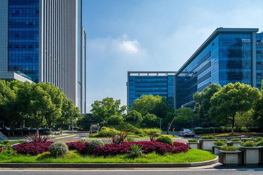 Office buildings with blue glass exteriors surround a landscaped median with lush greenery and flowers under a blue sky.