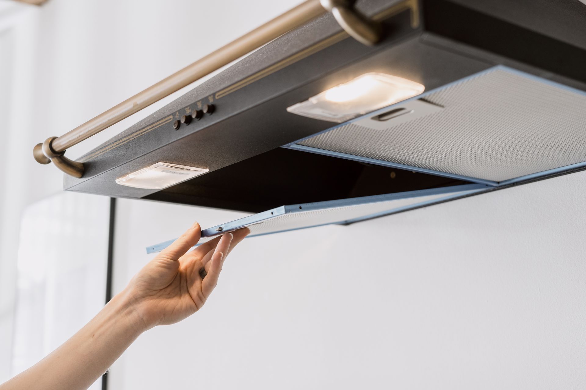 Hand opening a kitchen range hood vent to reveal a mesh filter and lights.