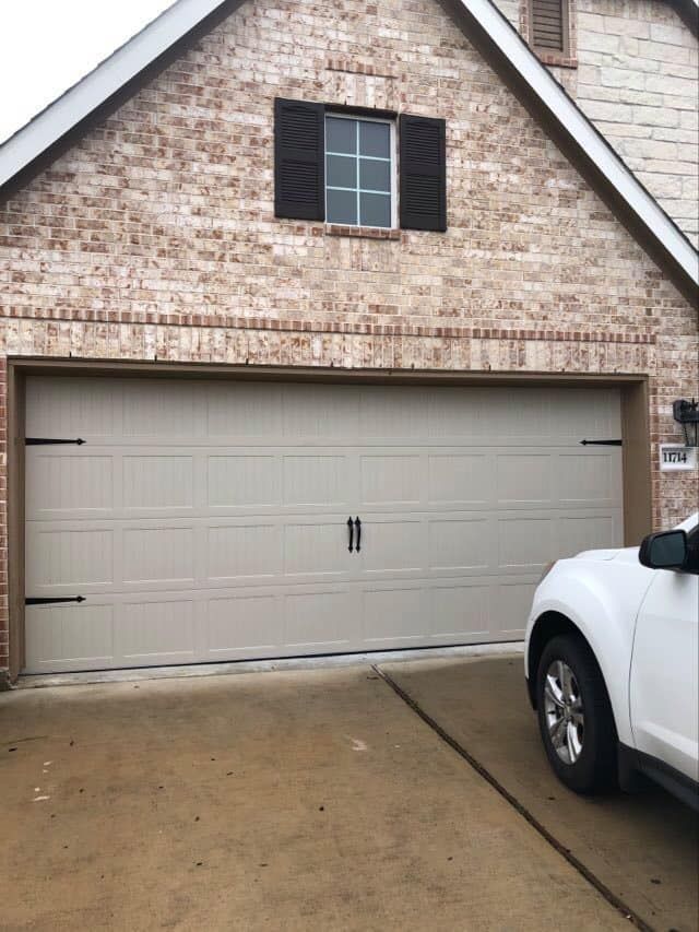 Tan garage door with black hardware on a brick house; a white car is parked nearby.