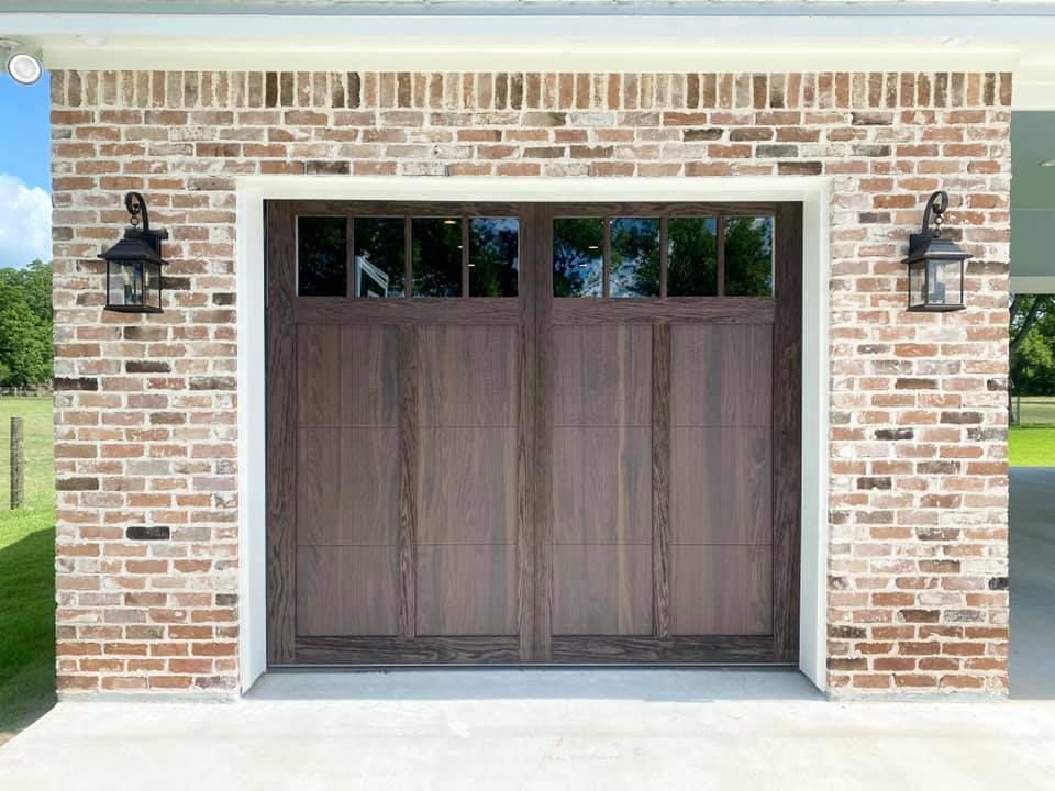 Brown wooden garage door with glass panes, flanked by brick, and lanterns.