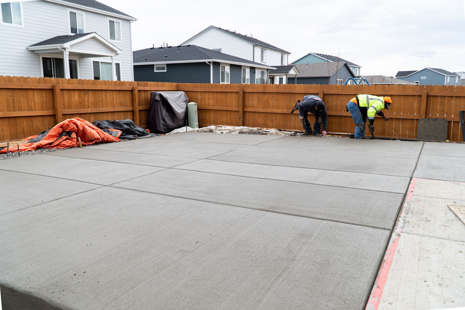Construction workers pouring concrete on a gravel surface. One worker guides the flow, another watches.