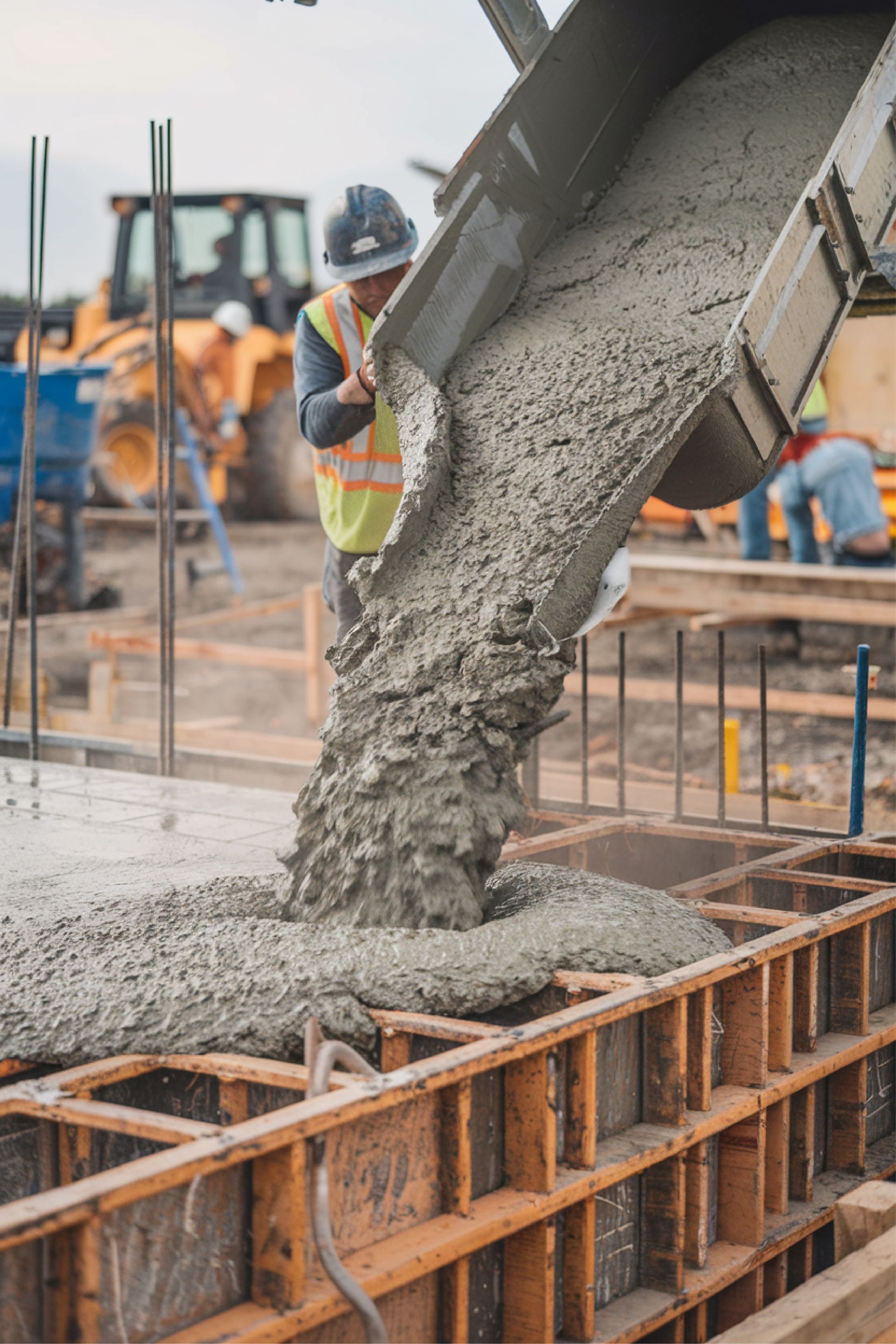 Concrete being poured into wooden forms at a construction site by a worker in safety gear.
