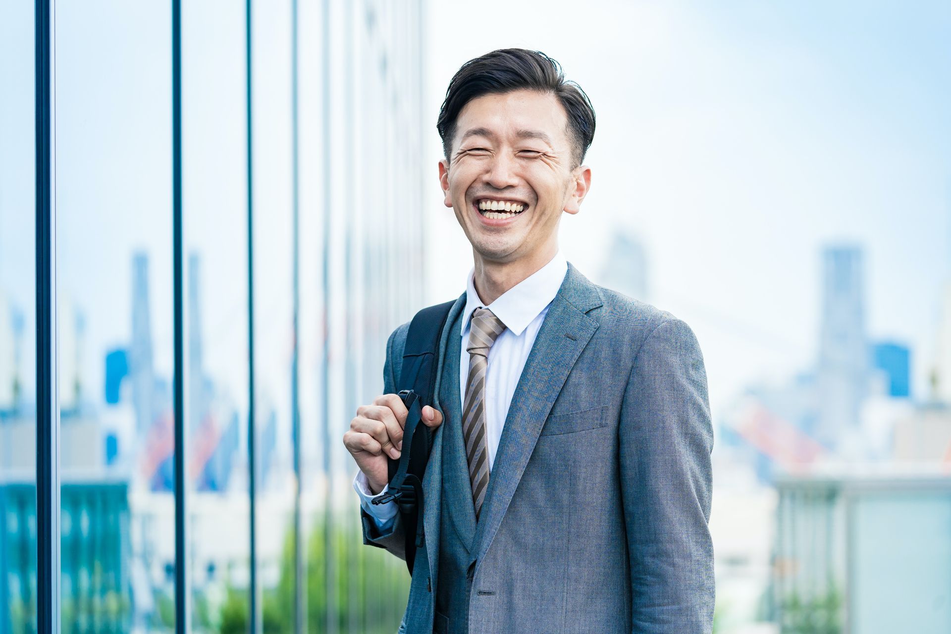 Smiling man in a gray suit holding a shoulder bag outdoors by a glass building.