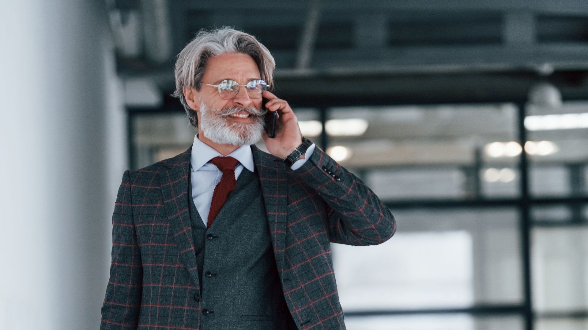 Man in a dark plaid suit talks on a phone in a modern office lobby.