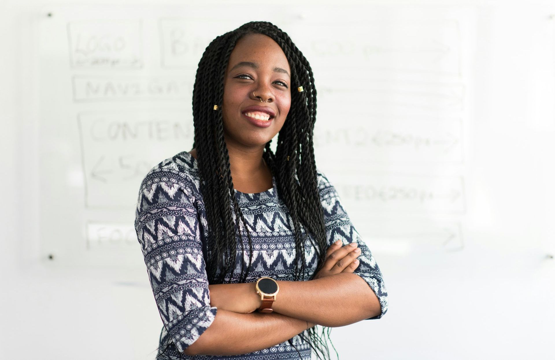 Woman with braided hair, arms crossed, smiling in front of a whiteboard.