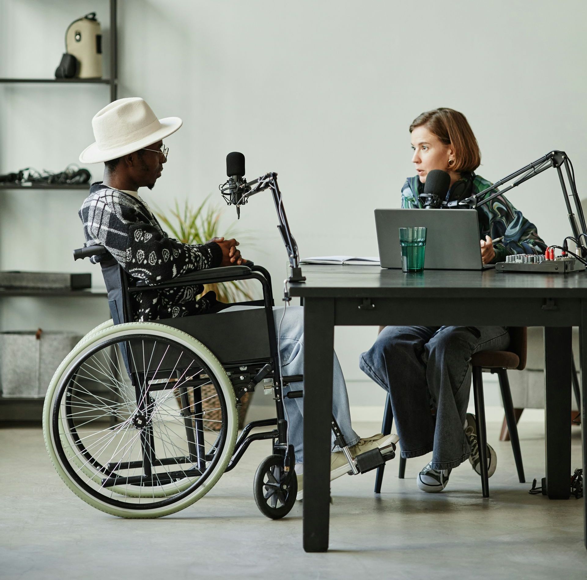 Man in wheelchair and woman at a table with microphones, podcast recording. Indoor setting.