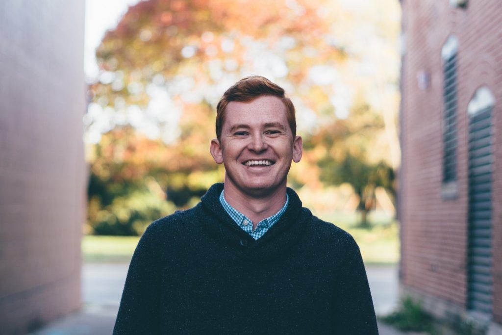 A man in a black sweater is smiling in front of a brick building.