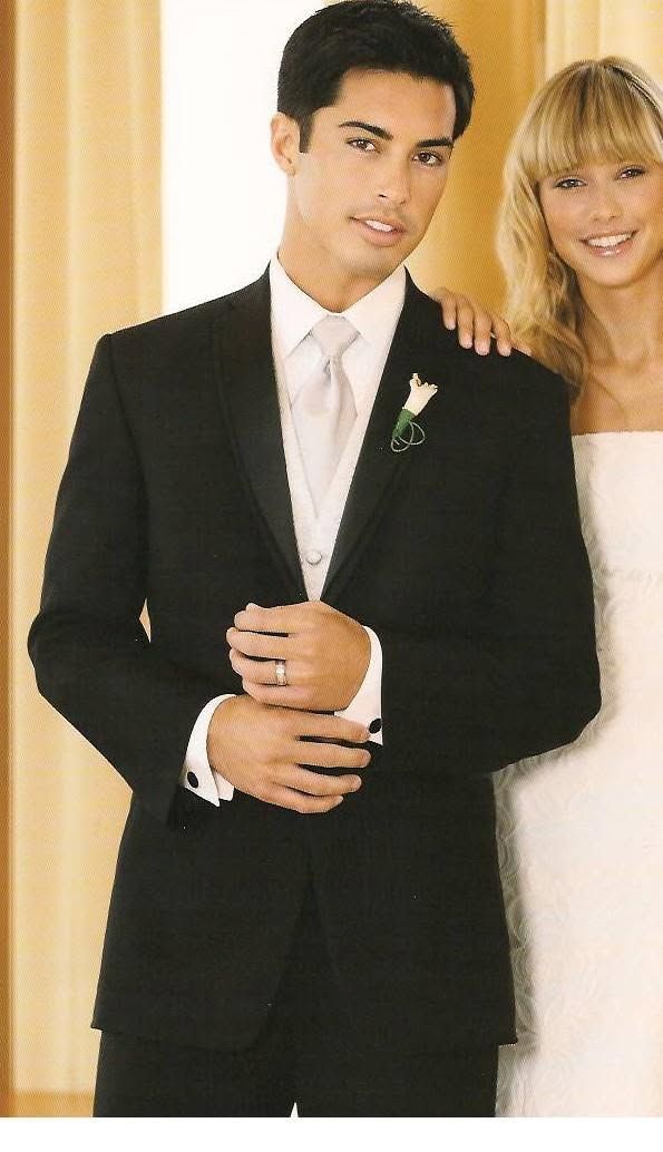 Man in a black suit with a white tie stands indoors, smiling beside a blonde woman in white.