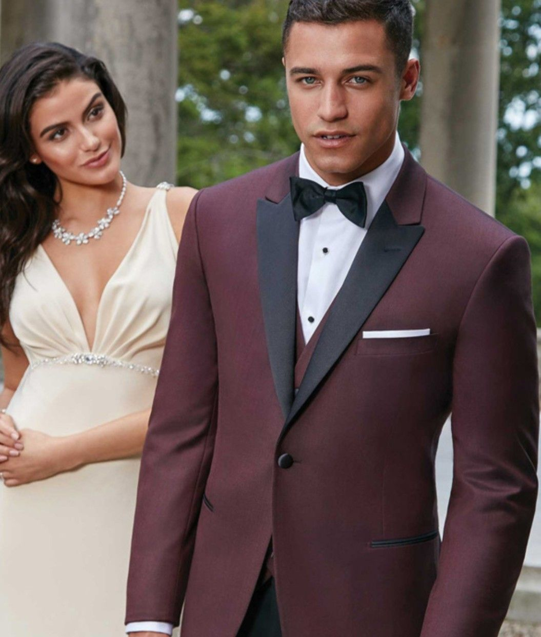 Couple in formalwear outdoors, man in burgundy tuxedo and woman in cream dress, posing for a photo