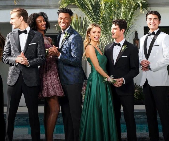 Five formally dressed people pose together in front of palm trees at a stylish evening event.