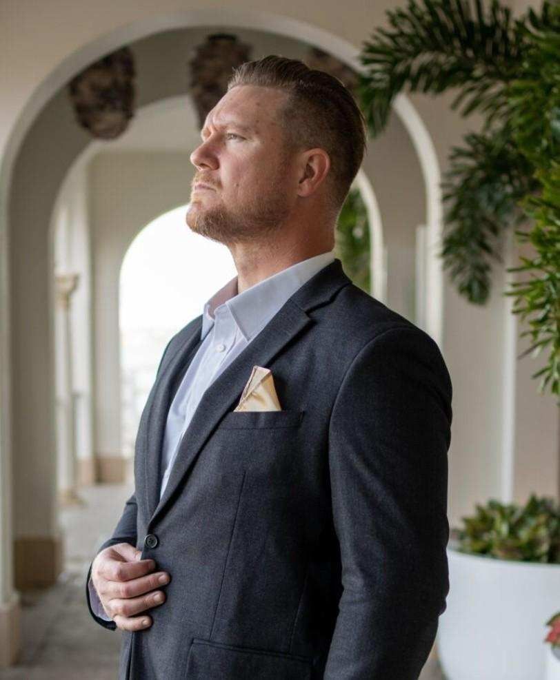 Man in a dark suit standing in an arched hallway, looking upward with a hand on his chest.
