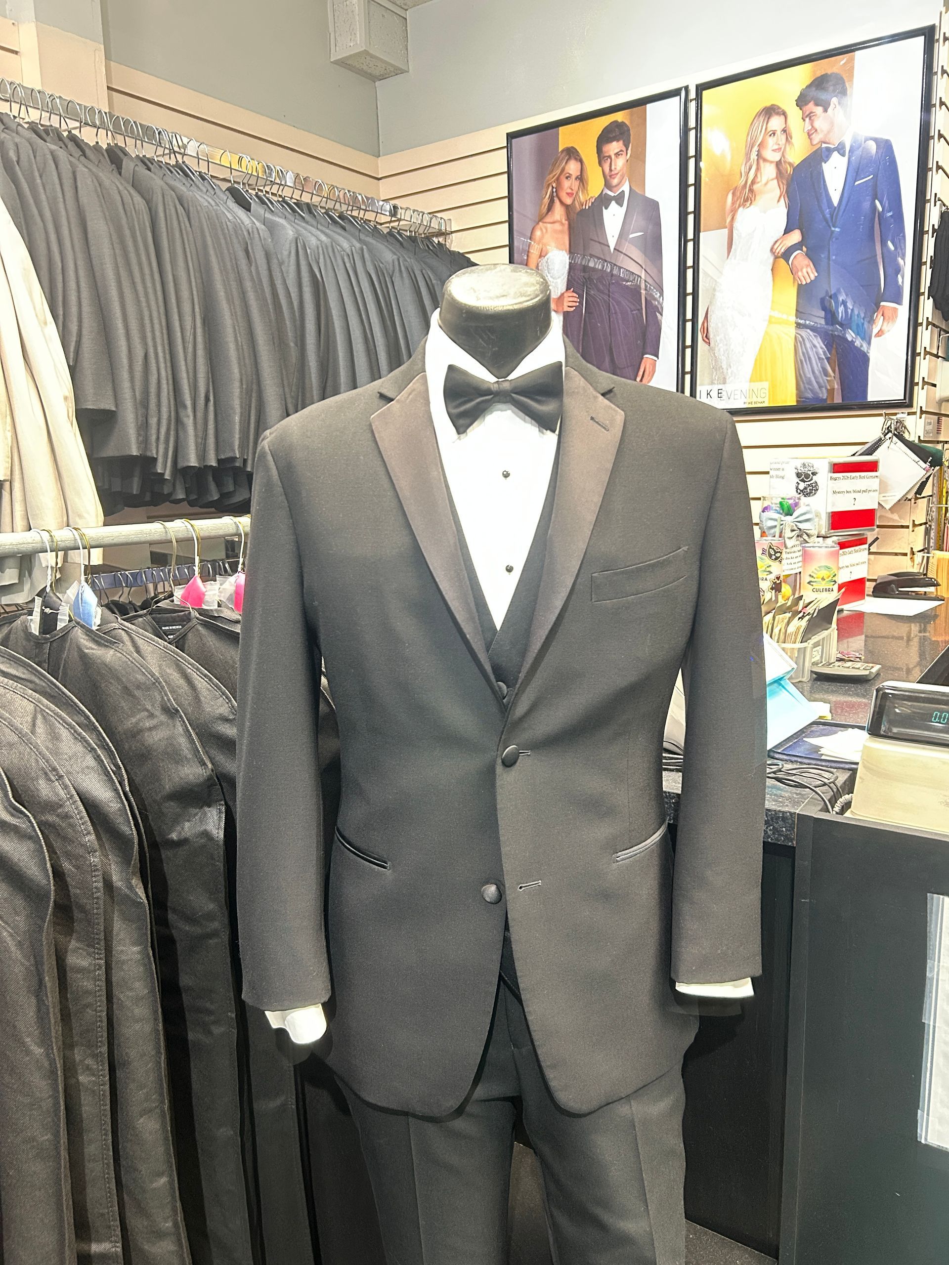 Black tuxedo suit on a mannequin in a clothing store, with gray jackets and posters in the background