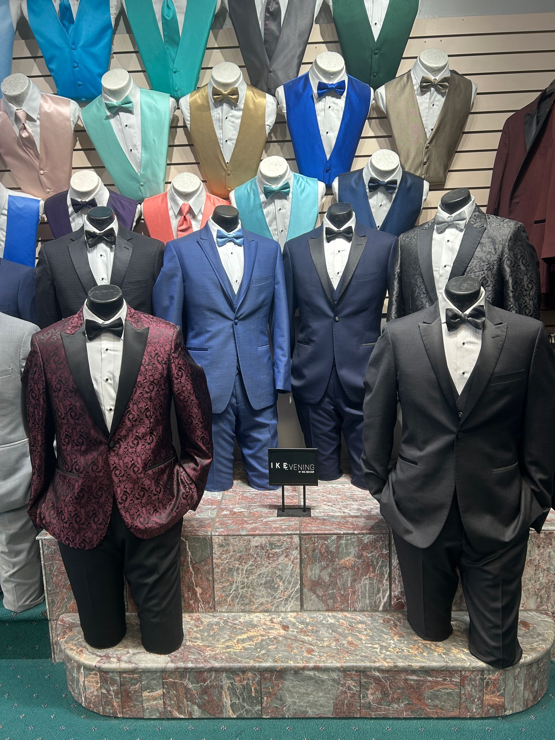 Mannequins in tuxedos and suits displayed on tiered steps in a formalwear store.