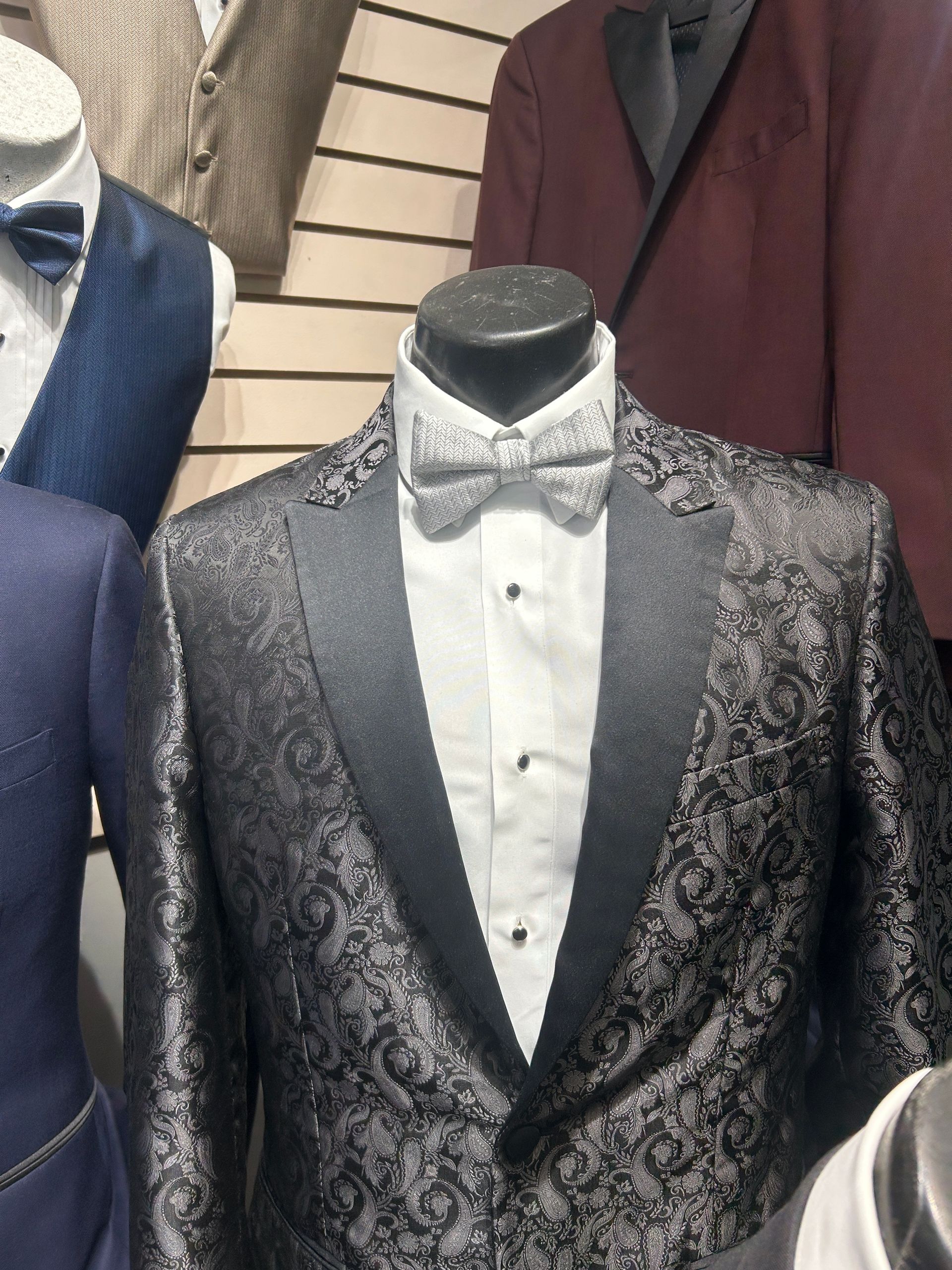 Black paisley tuxedo jacket with satin lapels on a mannequin, shown with a white dress shirt and bow tie.