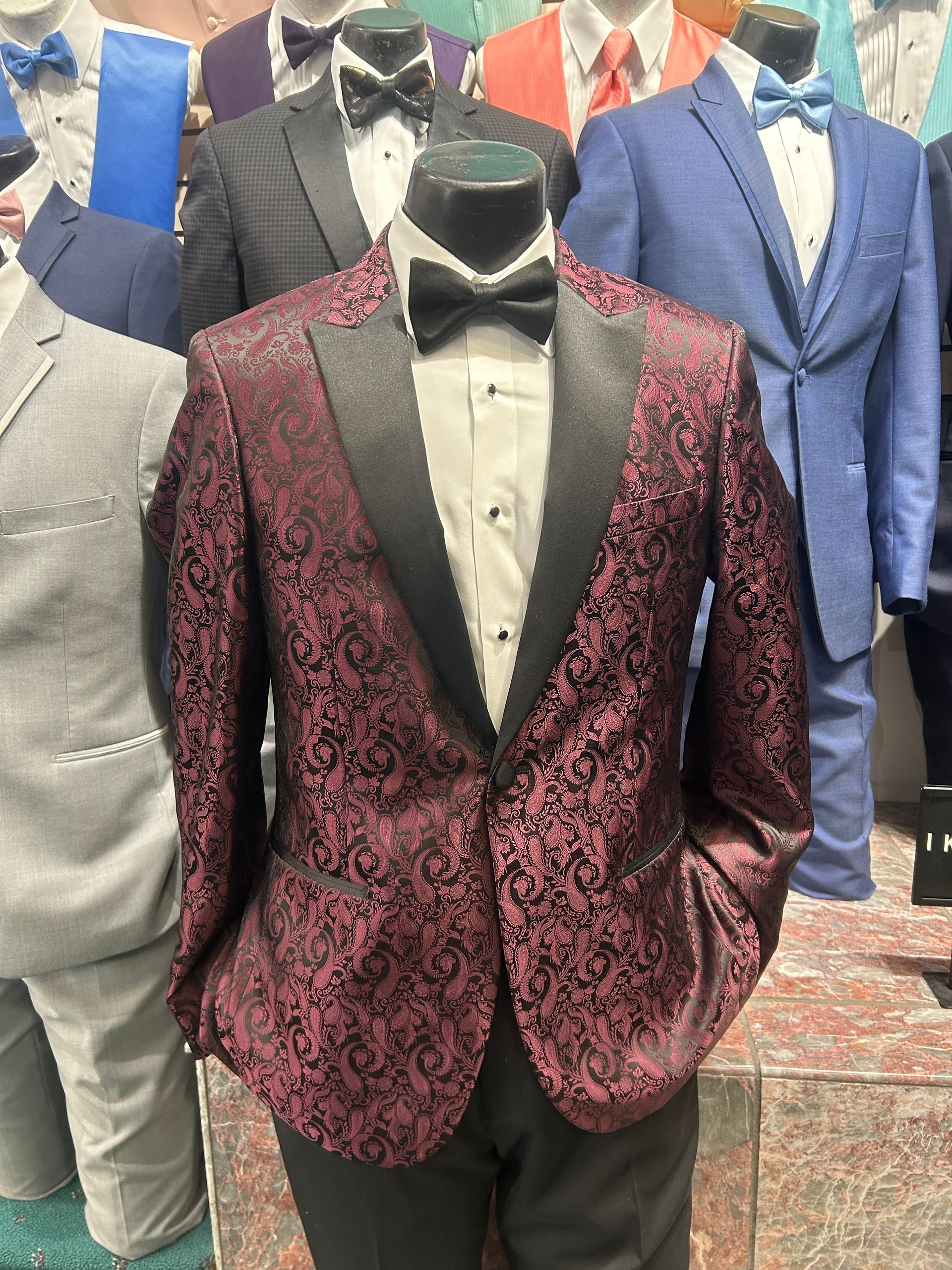 Mannequin in burgundy patterned tuxedo jacket with black lapels, shown among other suits in a clothing display
