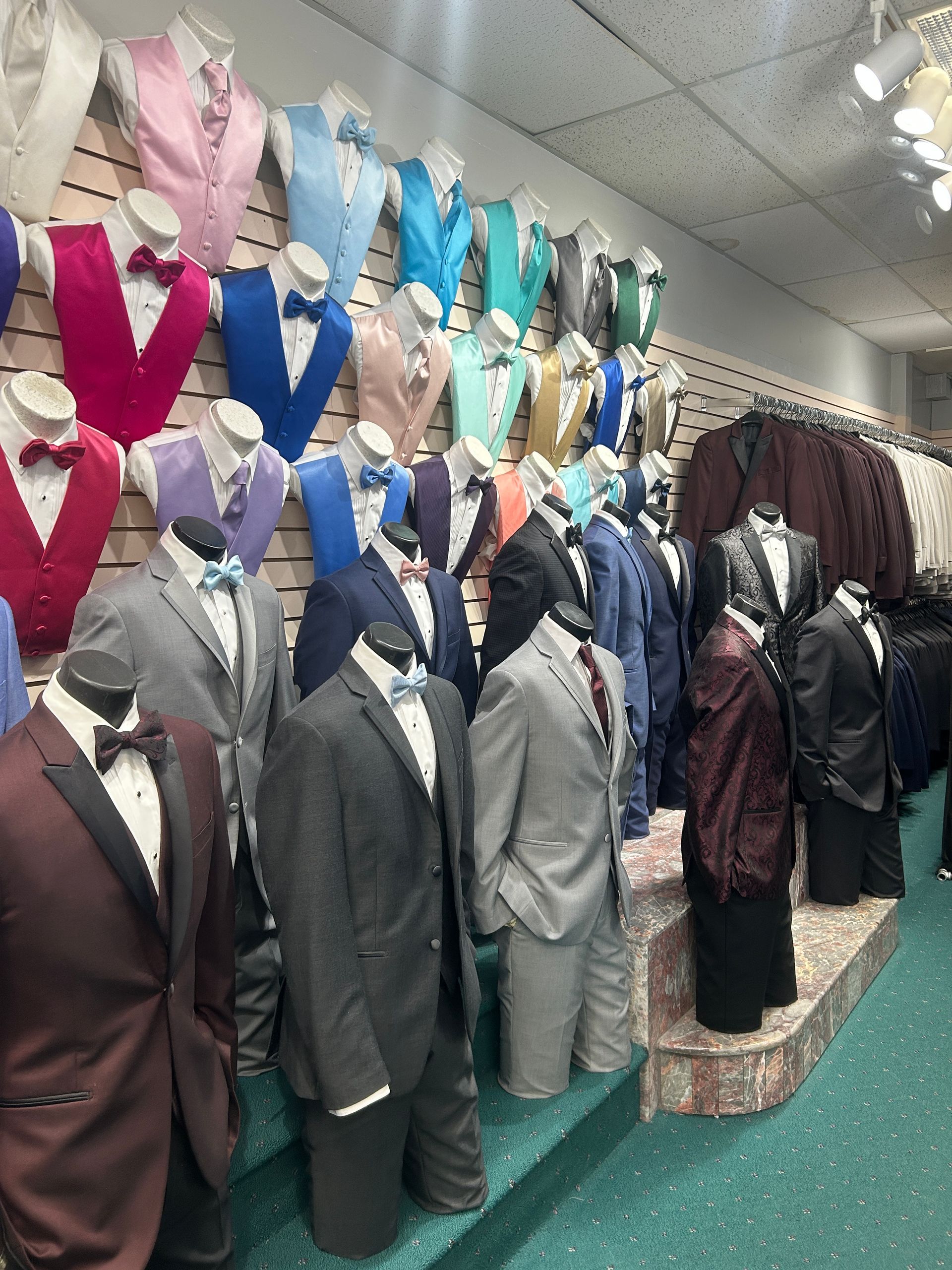 Rows of men’s suits and vests on mannequins in a formalwear shop, in assorted colors including gray, blue, and burgundy