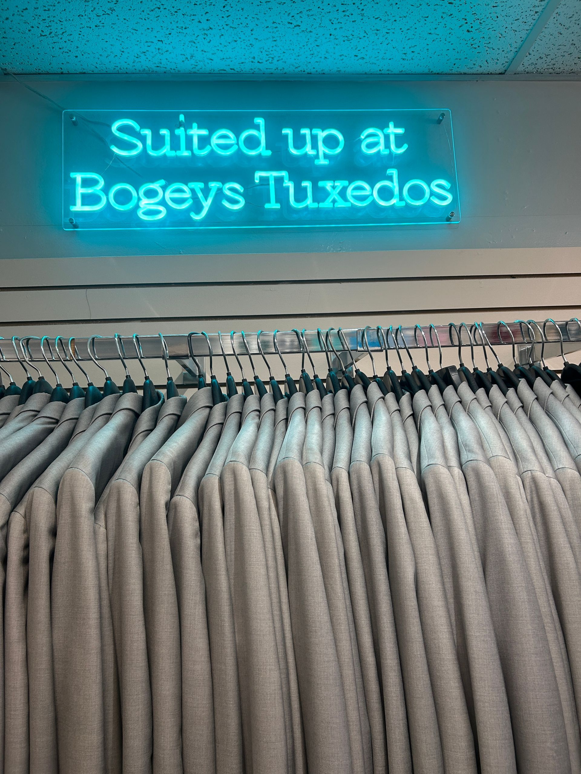 Neon sign reading “Suited up at Bogeys Tuxedos” above a rack of hanging suits