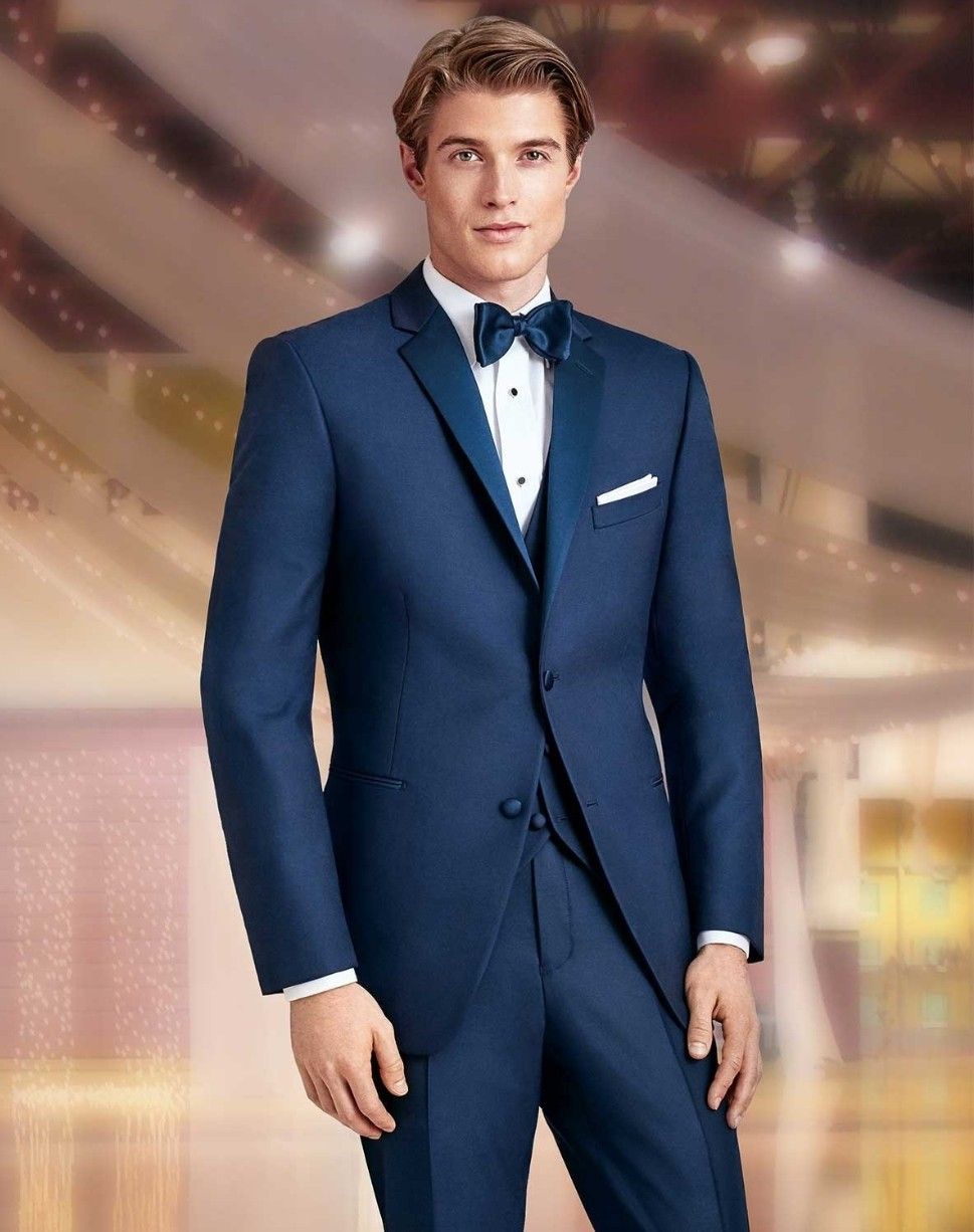Man in a navy tuxedo with a black bow tie, standing against a softly lit, elegant background.