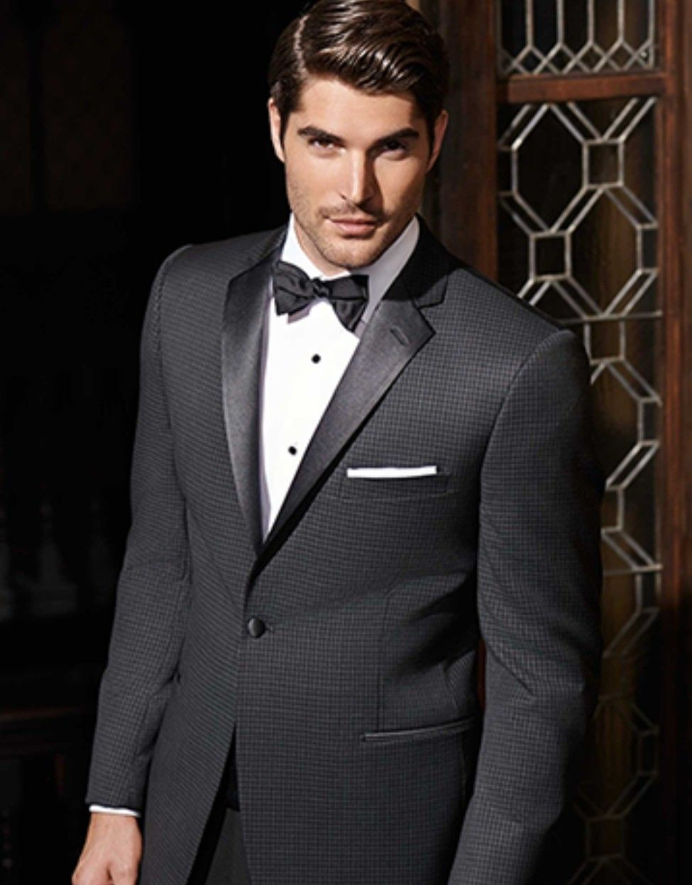 Man in a dark gray tuxedo with black bow tie, posing indoors by a decorative doorway.