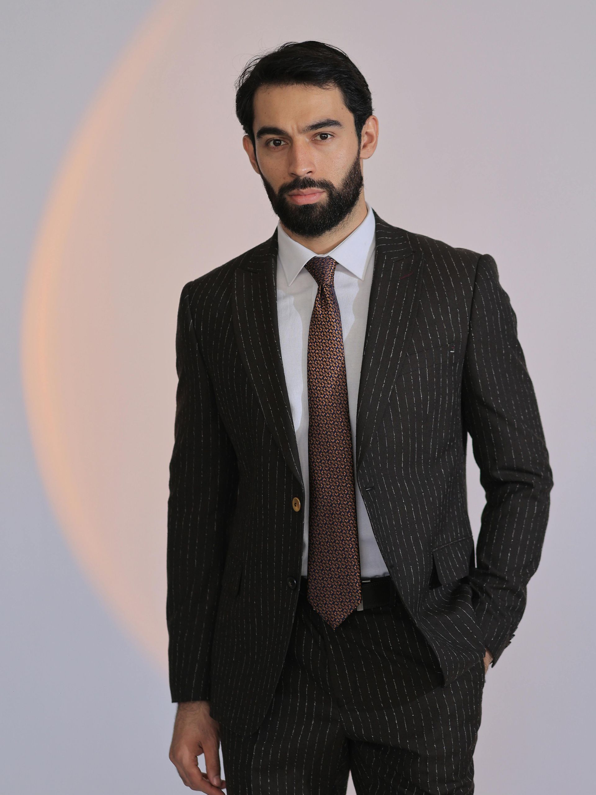 Man in a dark suit and patterned tie standing against a light background with a soft orange arc