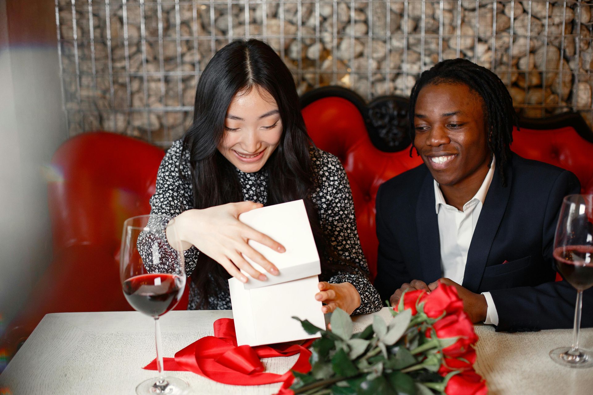 Two people opening a white gift box at a candlelit table with wine glasses and red roses