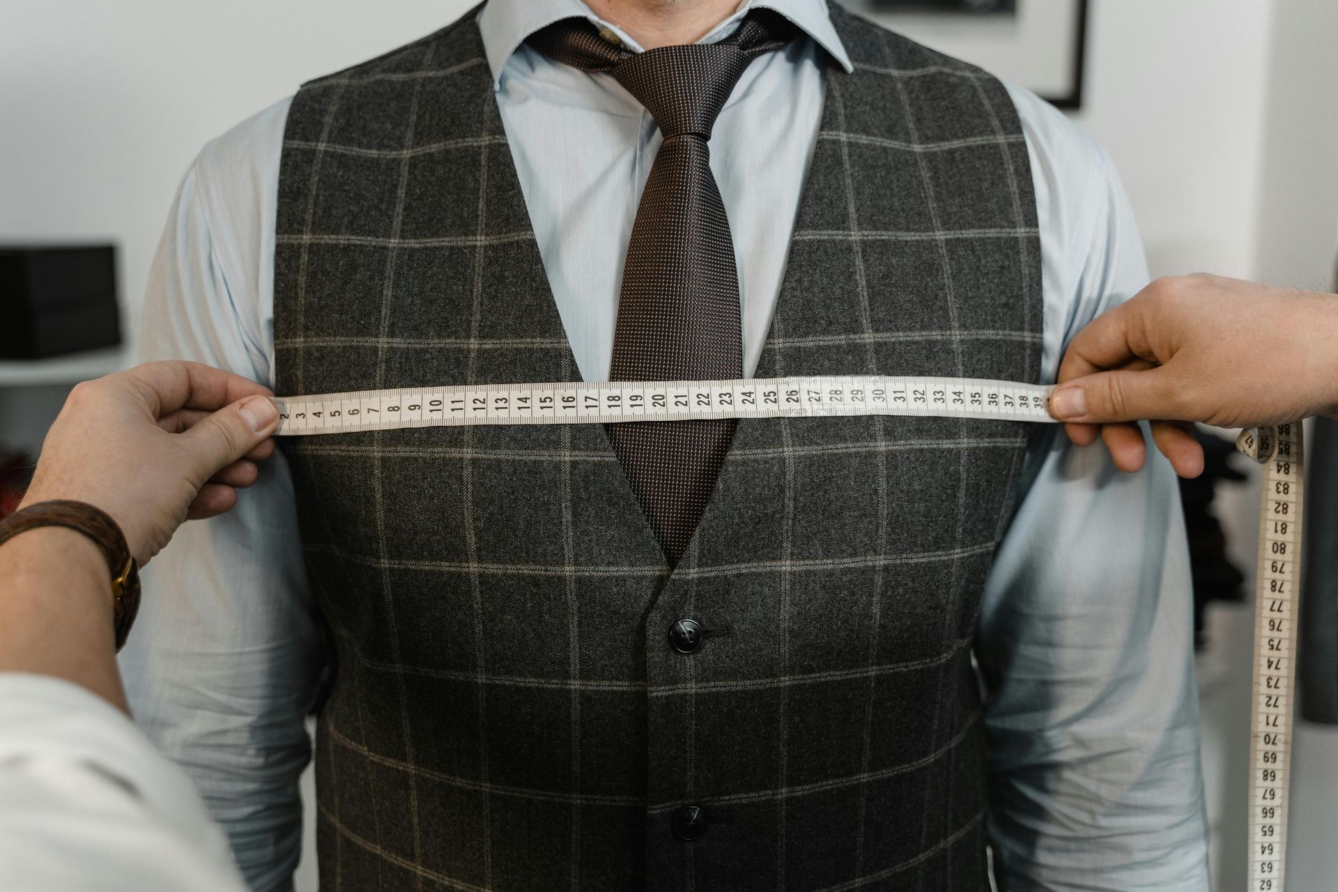 Tailor measuring a man's chest over a white shirt and dark plaid vest with a tape measure.