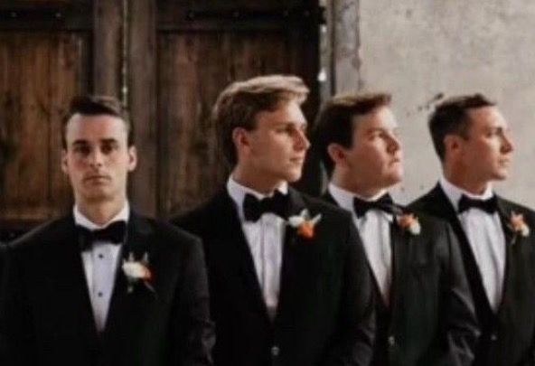 Four men in black tuxedos with boutonnieres standing in a row at a formal event