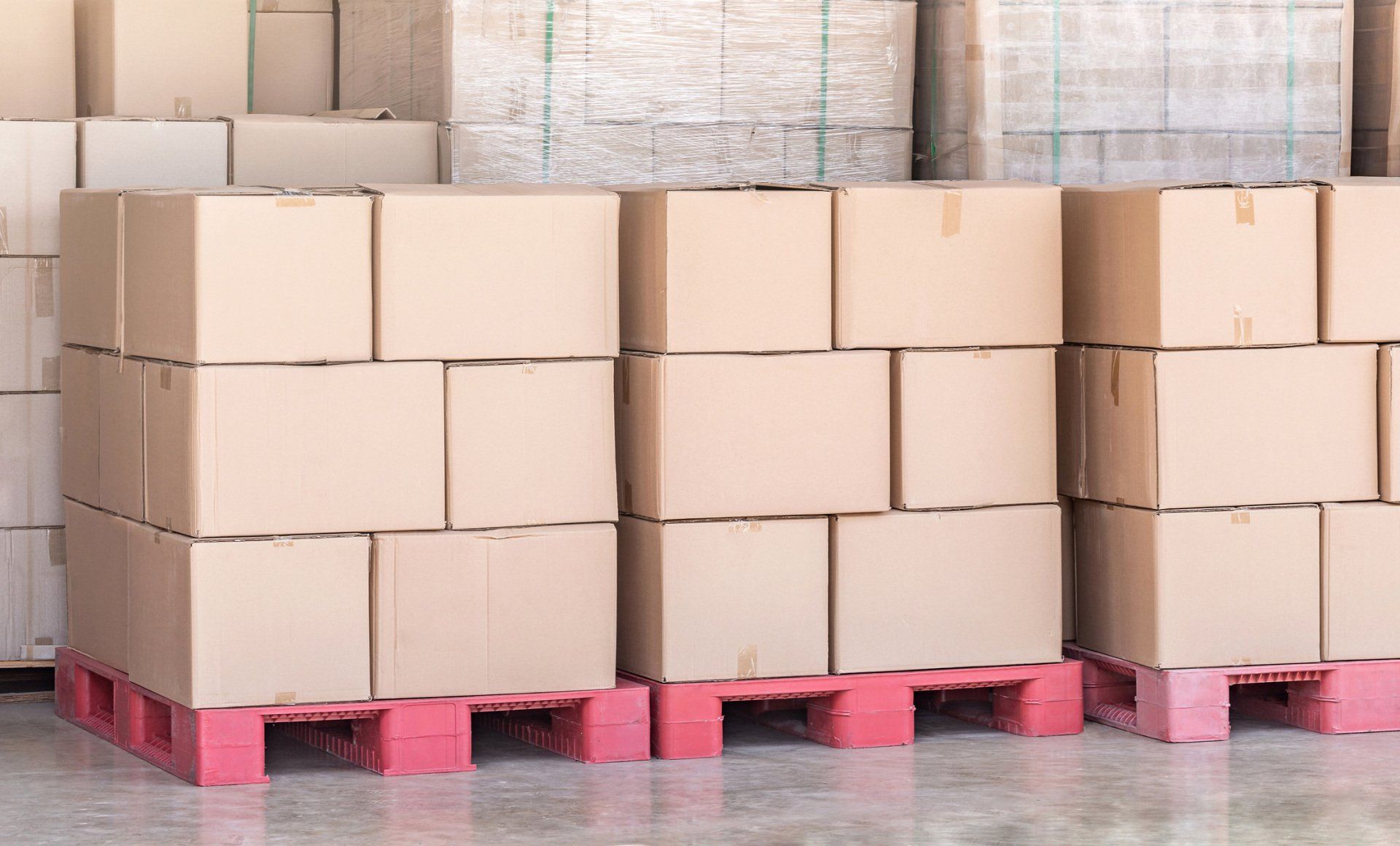 Stacks of brown cardboard boxes are organized on red wooden pallets in a warehouse.