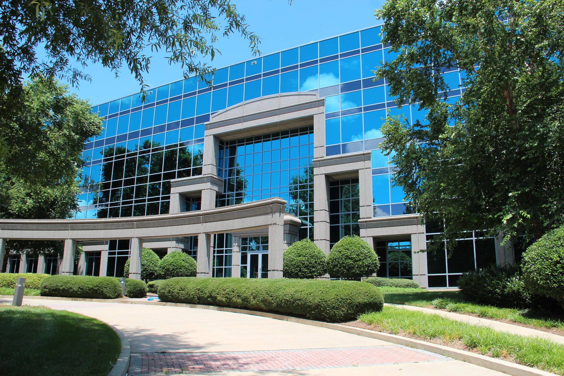 Modern glass office building with blue reflective windows and landscaped entrance on a sunny day