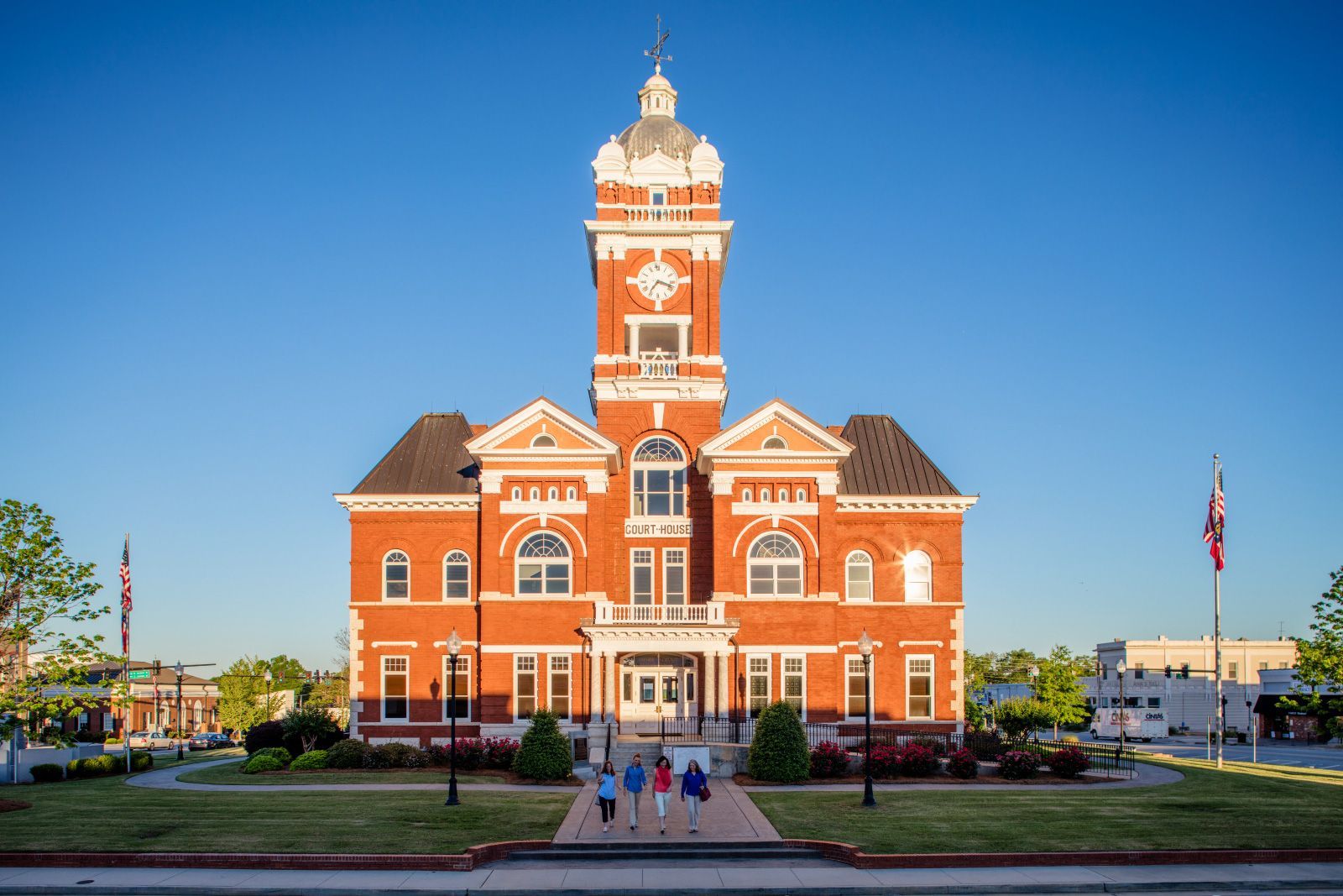 Red-brick historic courthouse with central clock tower under a clear blue sky, people on steps