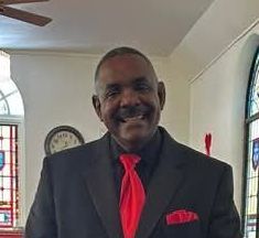 Man in a black suit with a red tie and pocket square, smiling, in a church.
