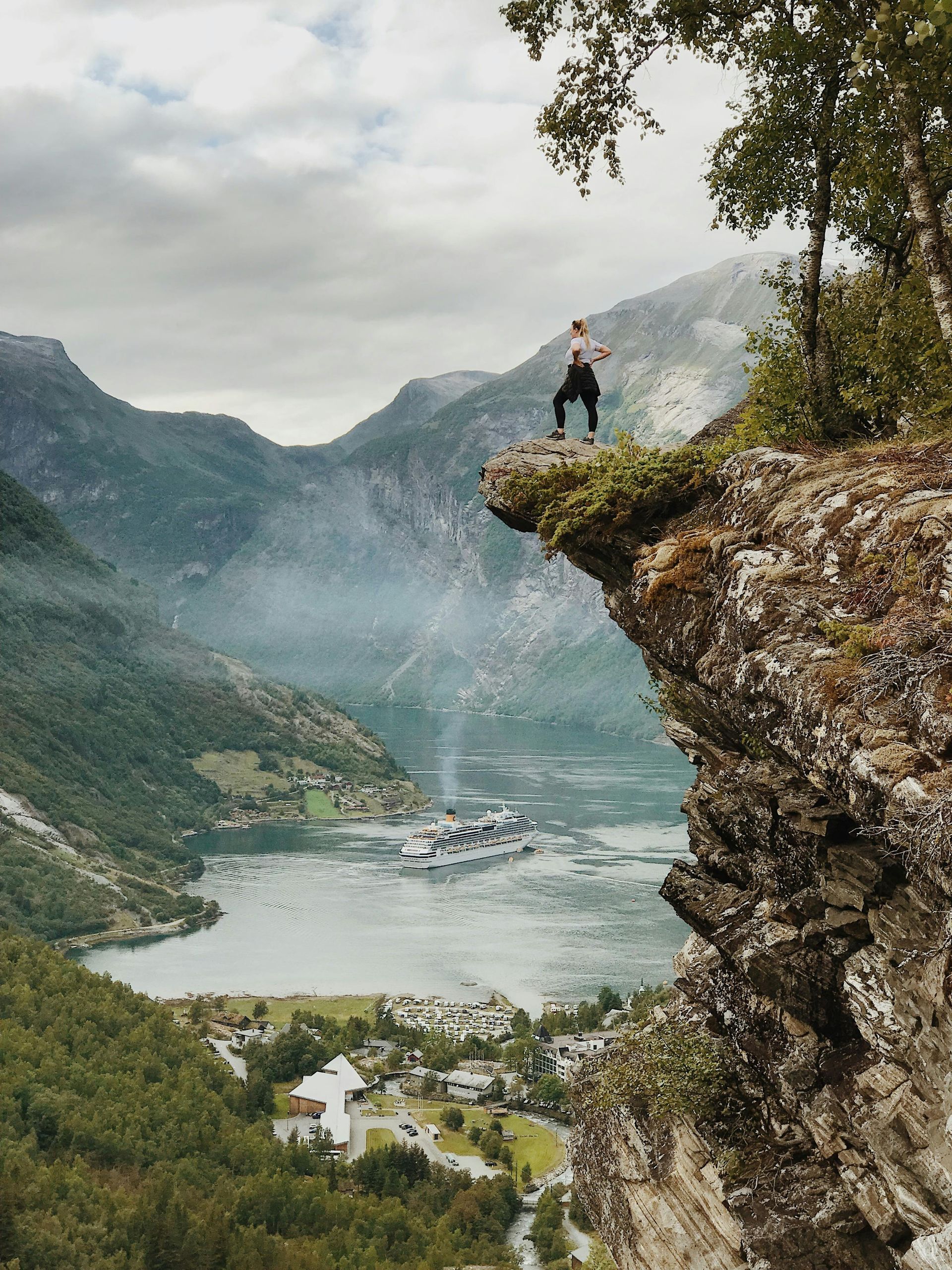 Person standing on cliff edge overlooking a fjord. Mountains and water in background. Cloudy sky.