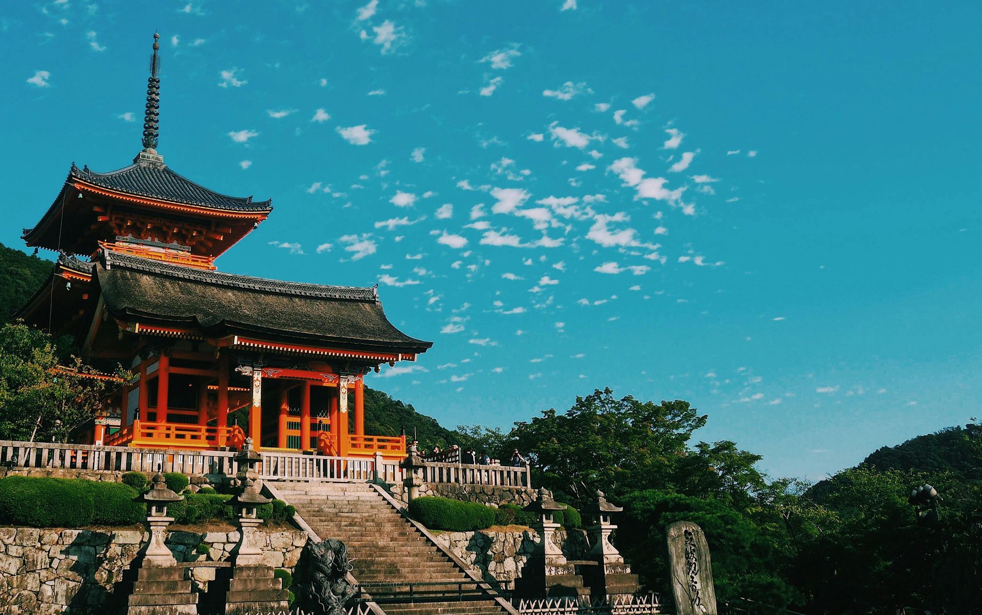 Japanese pagoda temple, orange and brown, under blue sky, lush greenery in the background.