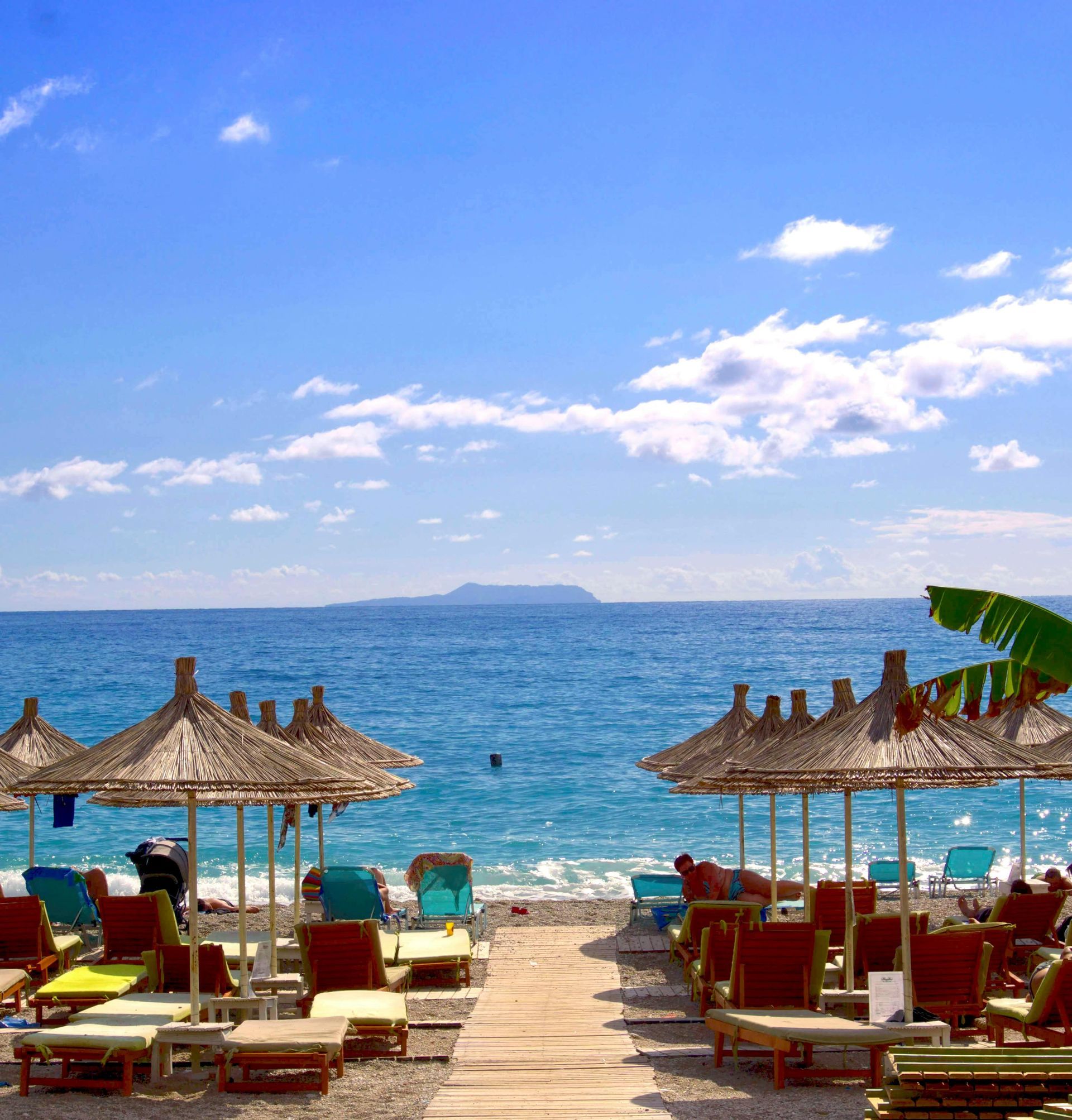 Beach scene with straw umbrellas, lounge chairs, and blue ocean under a sunny sky.