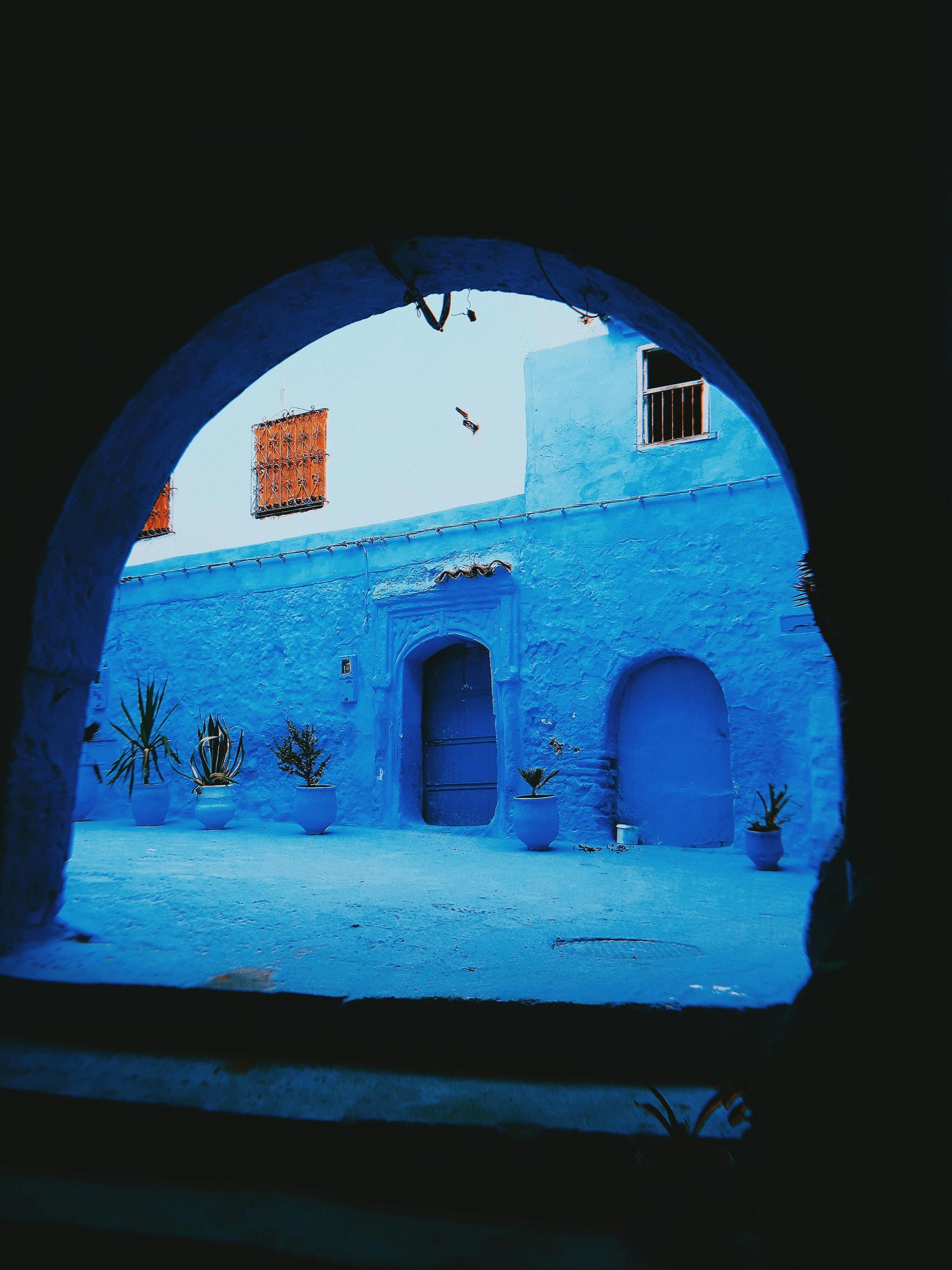 Arched doorway framing a vibrant blue courtyard with blue buildings, doors, and pots; terracotta shutters.