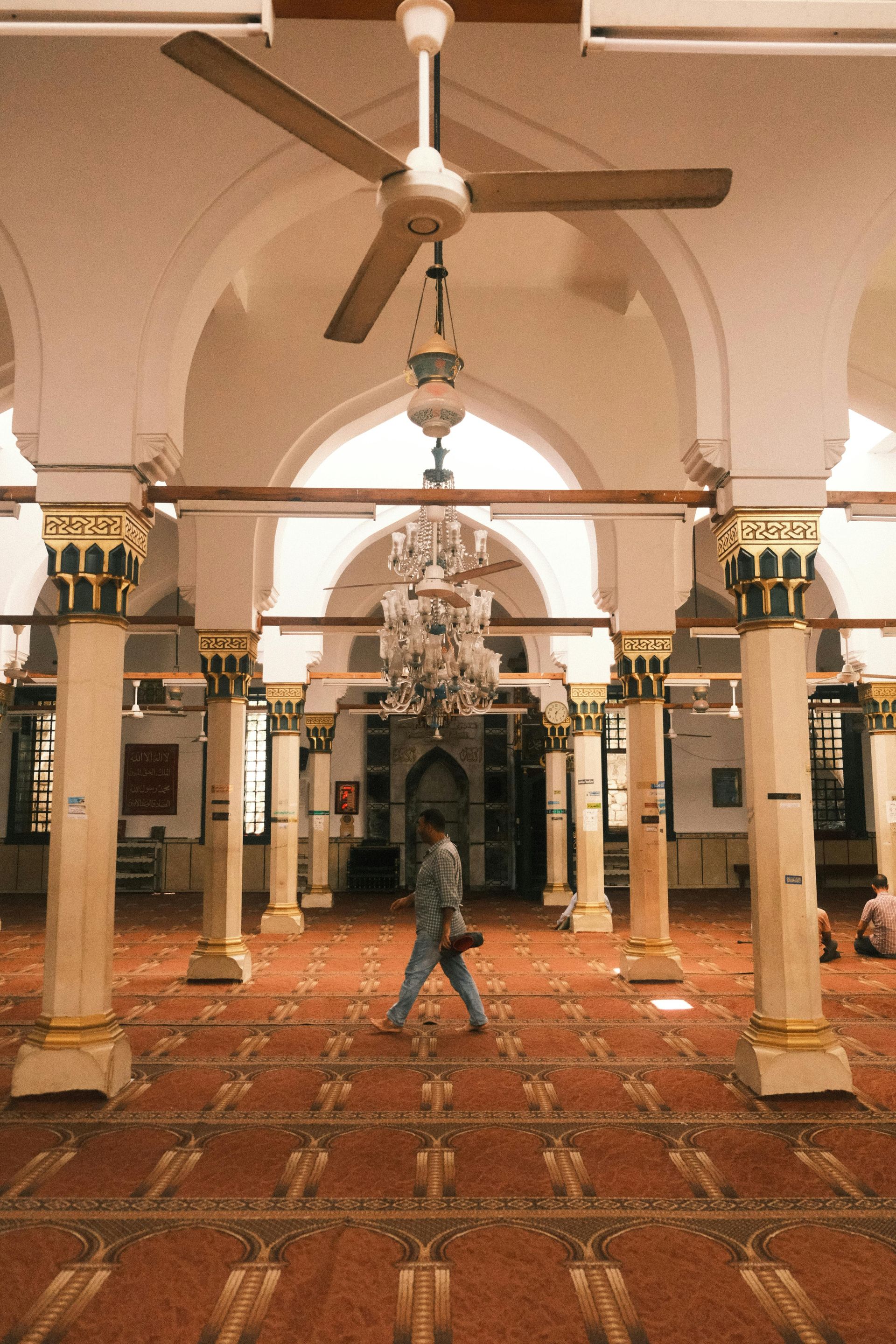Interior of a mosque with arched ceilings and pillars. A person walks on a red patterned carpet.