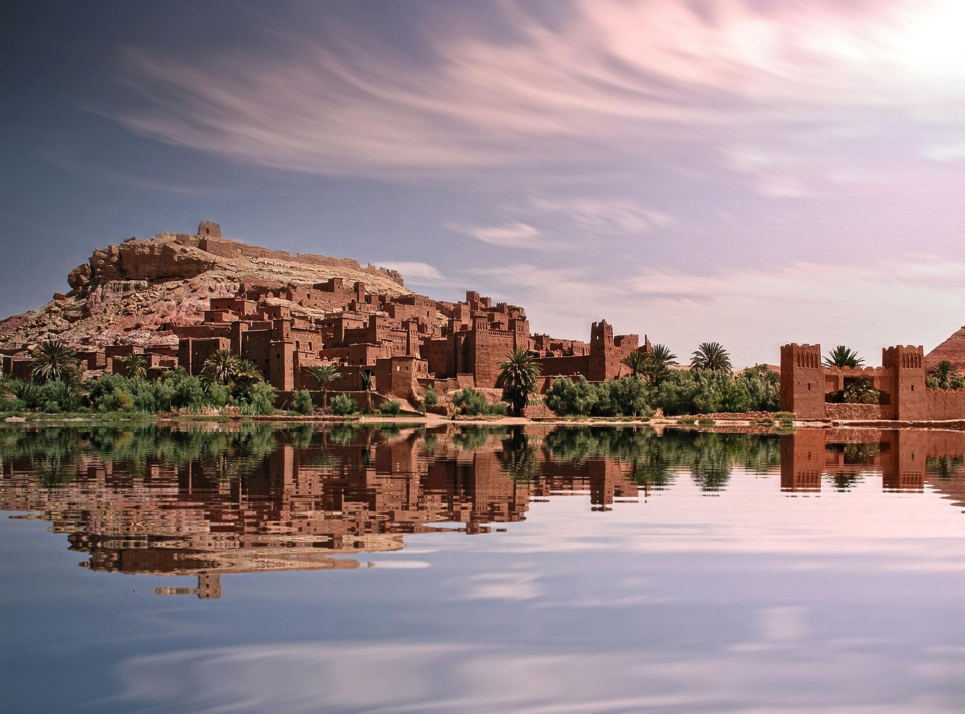 Ancient mud-brick town reflected in calm water, under a cloudy, pastel sky.