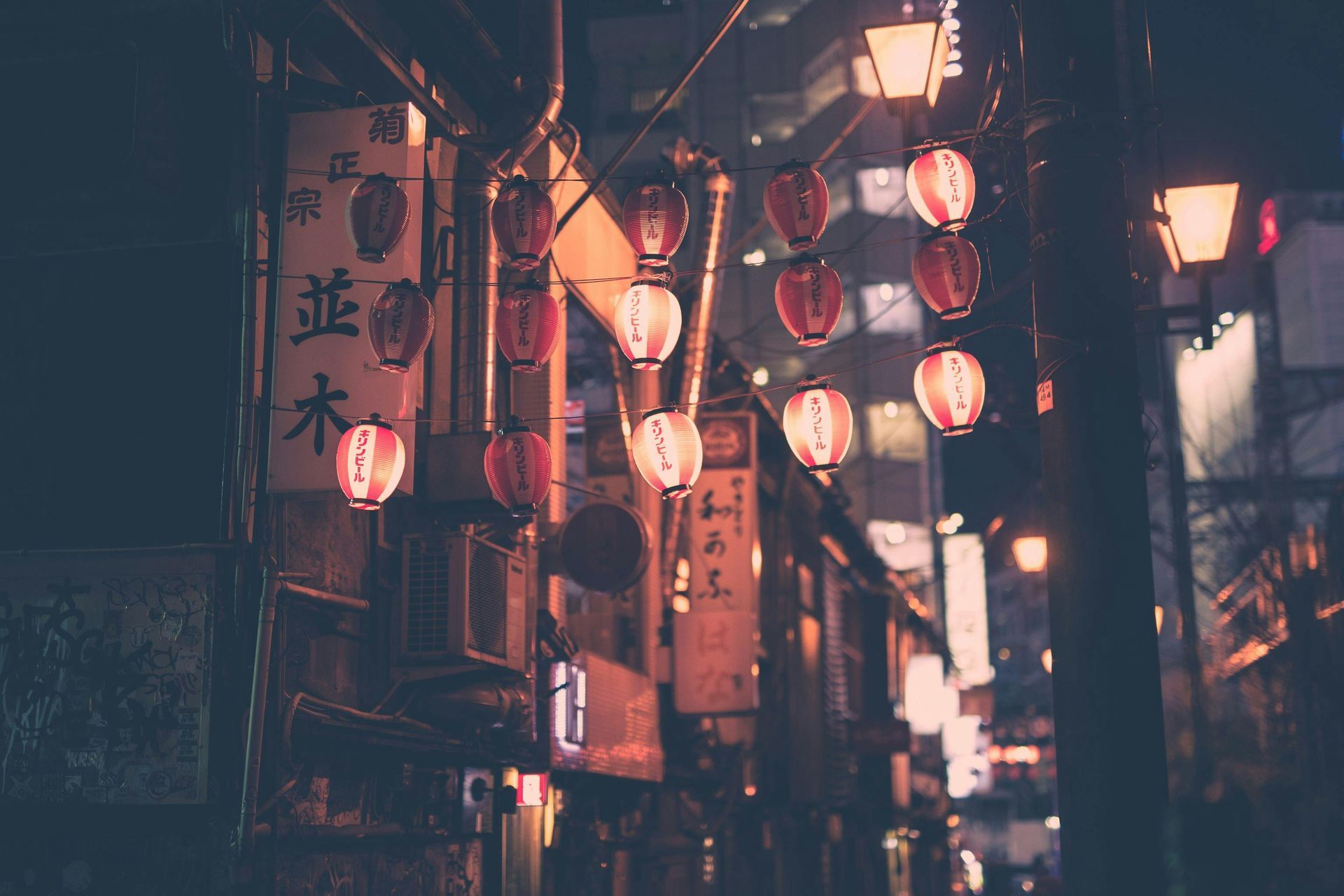 Night scene of a Japanese street with hanging red lanterns and shop signs, buildings in the background.