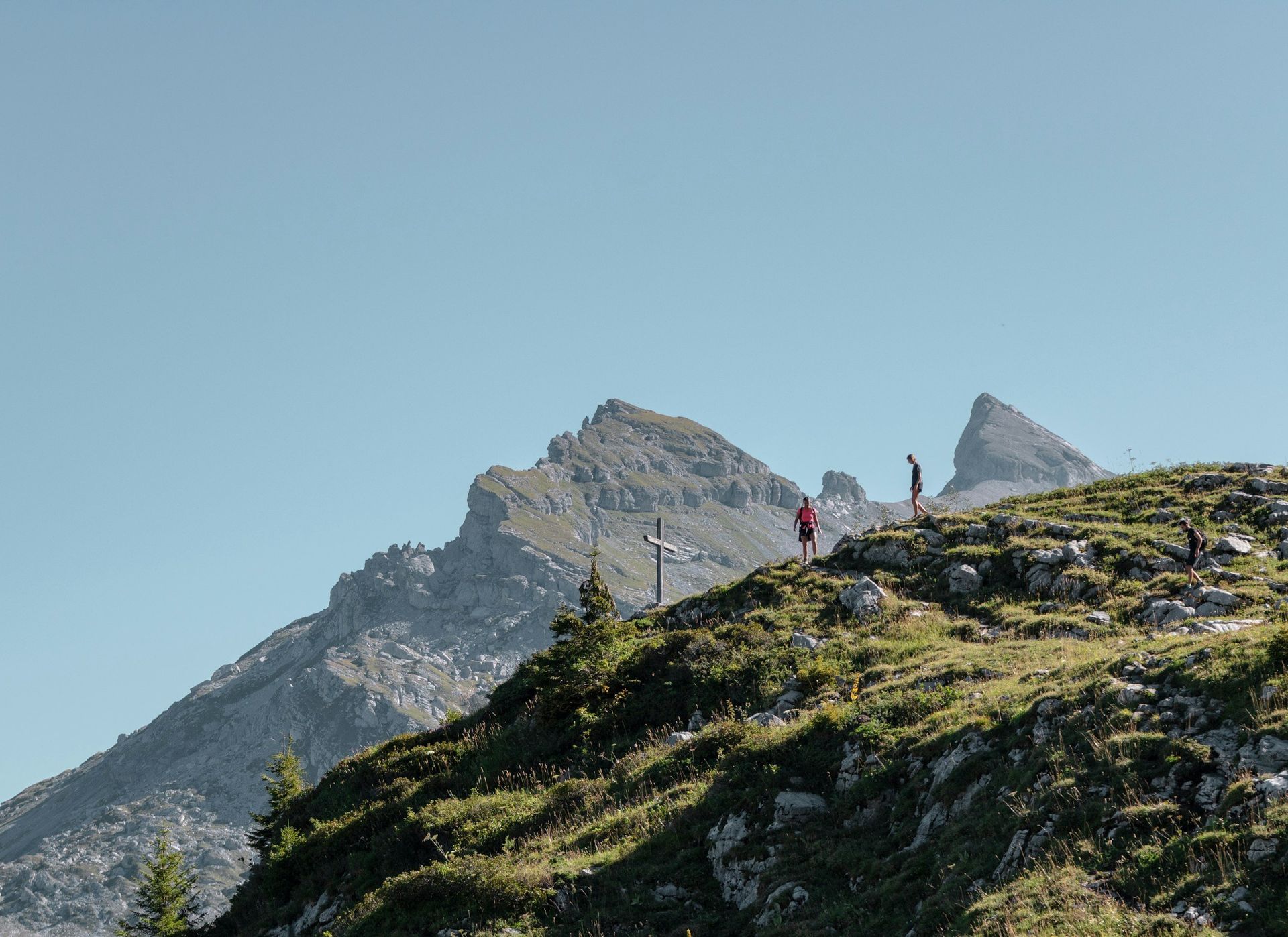Mountain hikers on a grassy slope with peaks against a clear blue sky.