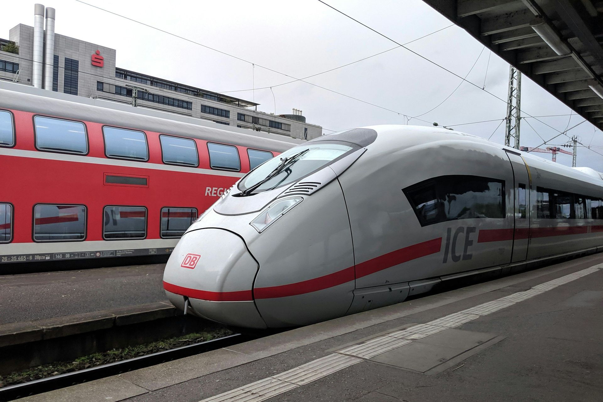 High-speed ICE train at a station platform, next to a red passenger train car, under a cloudy sky.